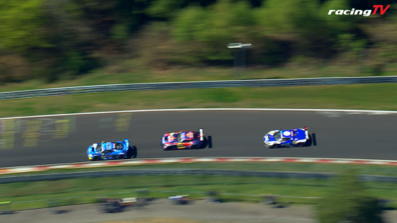 Three racing cars on a curved track in motion blur, blue and red livery, with RacingTV logo in the corner.