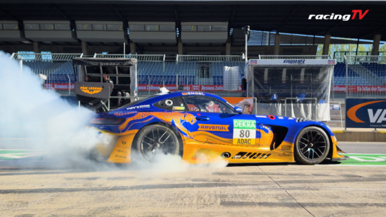 Blue and orange race car doing a burnout in the pit lane, smoke billowing from the tires, RacingTV logo visible in the top-right corner.