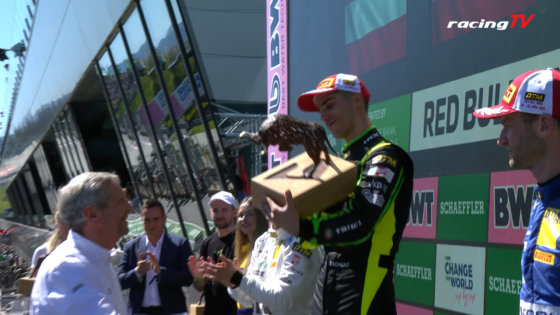 Race winner on the podium holds a wooden trophy box while a crowd applauds; racingTV logo in the corner.