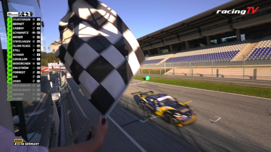 Person waving a large checkered flag beside a race track as cars pass; scoreboard overlay on the left and a RacingTV logo in the top-right corner.