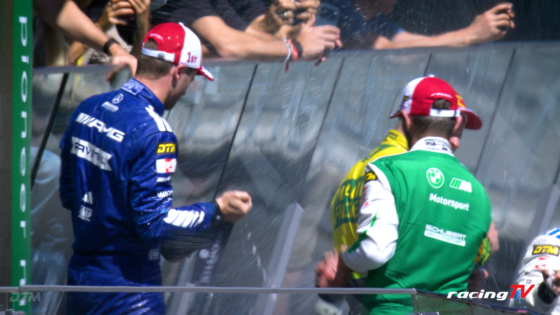 Two race crew members in blue and green uniforms stand at a barriers, with fans reaching over the railing behind them.