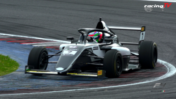 Silver open-wheel race car with #7 on the nose speeding through a blue-curbed corner, driver wearing a bright multicolor helmet; Racing TV logo in corner.