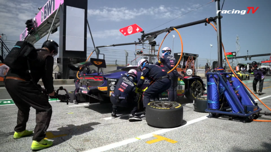 Pit stop at a race track: crew changing a wheel on a race car while a photographer films nearby.