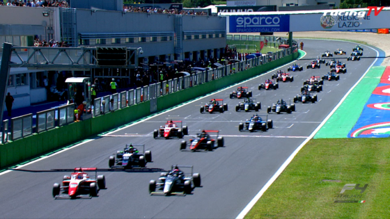 Multiple Formula 1 cars race down a track toward the camera, with a pit complex and spectators along the wall.