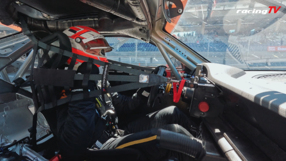 Driver in a red-and-white helmet strapped into a race car cockpit with a full harness and roll cage visible, windshield and controls in view