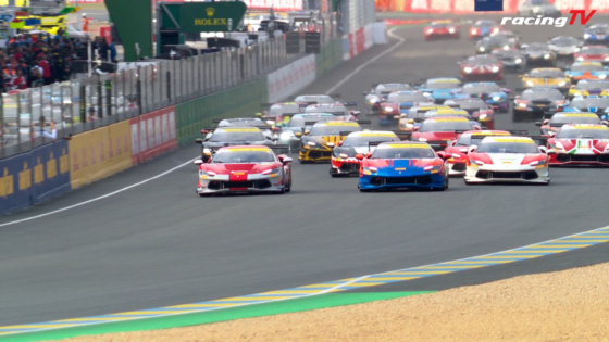 Multiple colorful race cars speeding down a track, with barriers and pit area on the left and a racingTV watermark in the corner.