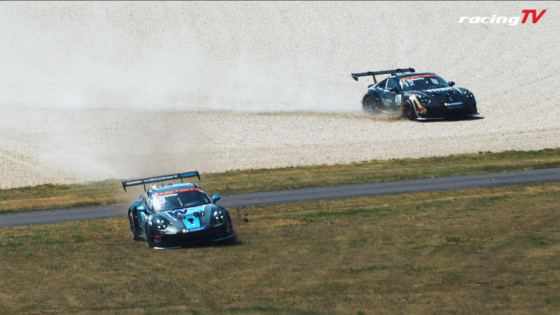 Two race cars speed around a gravel track, kicking up dust with the RacingTV logo in the top-right corner.