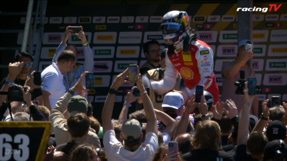 Motorcycle racer in red and white suit on a stage, fans grab smartphones to photograph him, sponsor boards in the background