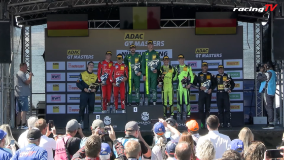 Podium ceremony at ADAC GT Masters with multiple racers in colored suits holding trophies, flags and sponsor banners in the background.