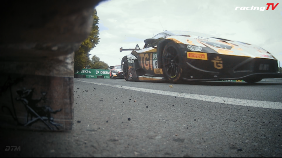 Low-angle shot of a black-and-orange race car on track, with green Dekra barriers in the background and racingTV logo in the corner.