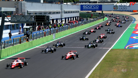 Formula 1–style race start with many open-wheel cars sprinting down the track past the pit building and green barriers, spectators watching above.