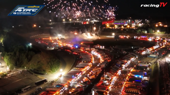 Aerial night scene of a truck race festival with rows of brightly lit trucks and fireworks above, logo banners in corners.