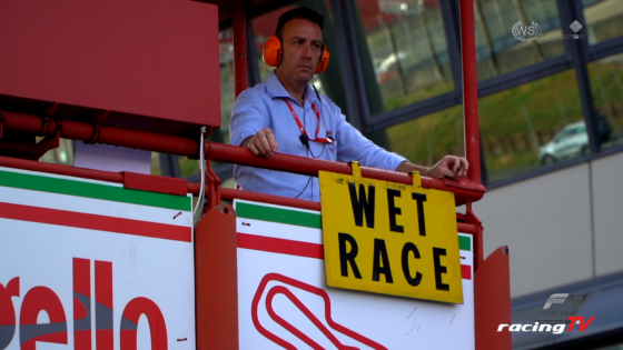 Race official in a blue shirt, orange headset, standing at a control platform with a yellow 'WET RACE' sign and sponsor banners nearby