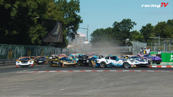 Multiple race cars crash at a corner, dust rising as crews watch the incident.