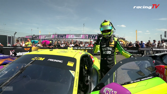Race car driver in neon green suit and helmet standing beside a bright yellow race car, with a crowd and banners in the background at a track festival.