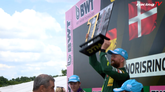 Winner on the podium lifts a gold trophy as fans cheer, with sponsor banners and a RacingTV logo in the background.