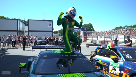 Race car driver in green suit stands on a car, fists raised in victory, as a crowd cheers in the pit area at a racetrack.