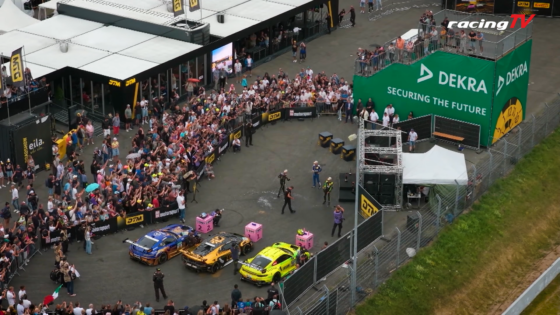 Aerial view of a crowded motorsport event with colorful race cars lined up near barriers and a green DEKRA wall in the background.