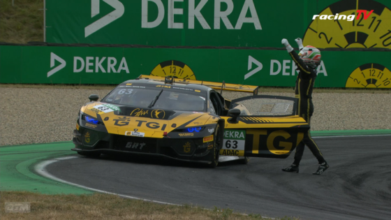 Race car #63 in yellow and black livery stops on a track as a crew member raises arms in celebration beside green Dekra barriers