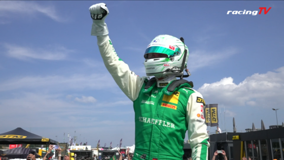 Race car driver in green suit raises fist in victory gesture, helmet on, a crowd cheers below with banners and tents.